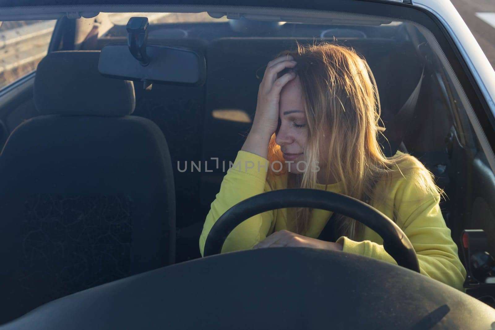 a girl of European appearance with blond hair in a yellow sweater sits behind the wheel of a car, sad, holds her head, does not know what to do, the car broke down on the road. High quality photo