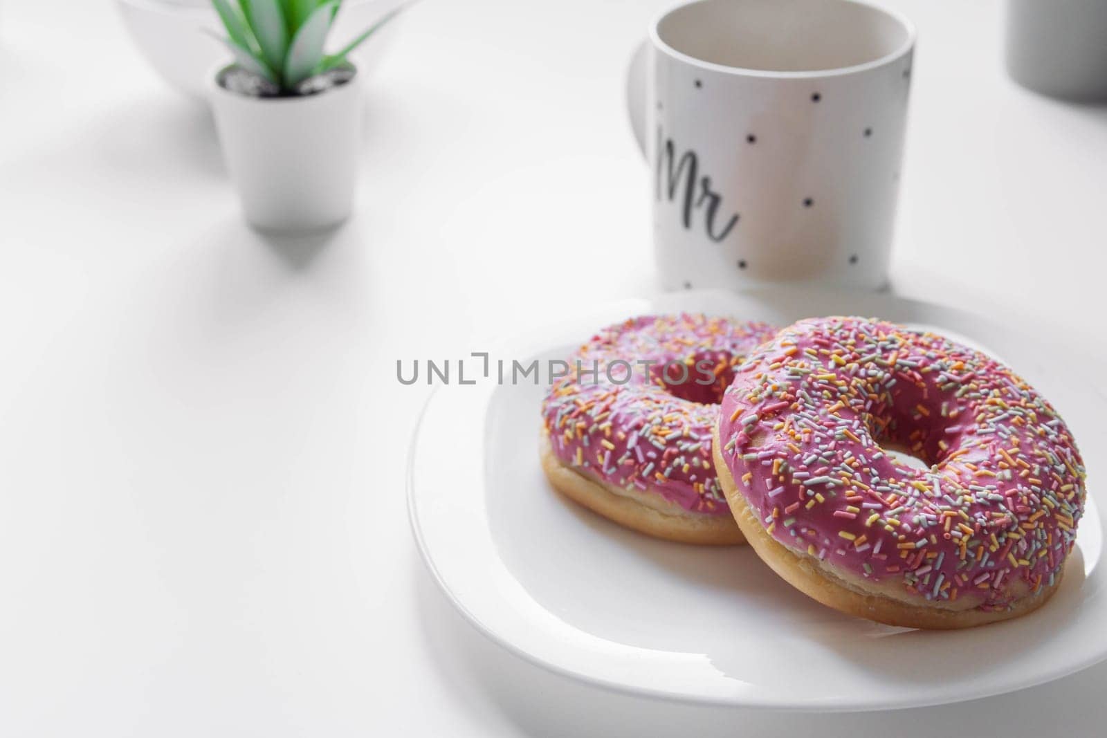 donuts with a pink icing and colored sprinkles on a white plate lie on a white table, next to a white cup with black dots. High quality photo on the left there is a place for the inscription