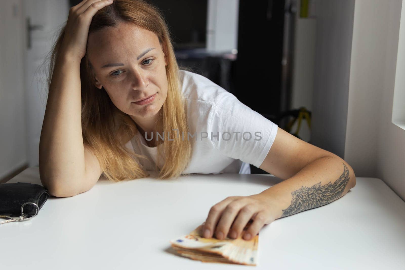 a girl of European appearance in a white t-shirt sits indoors at a white table with her head on her hands and the table is sad on the left hand of the girl with a tattoo, money lies nearby. by PopOff