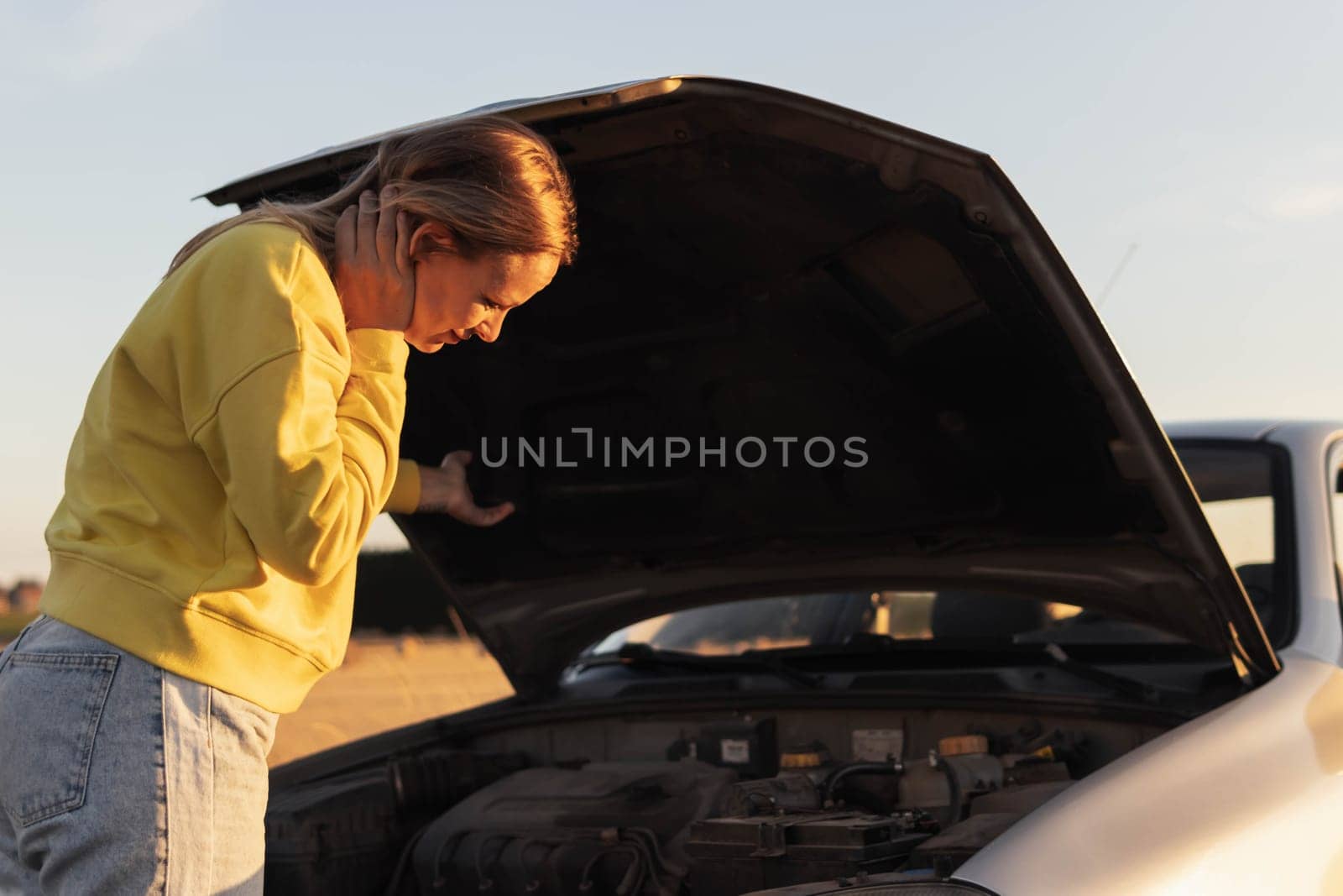 a girl with blond hair, stands with the hood of the car open and looks at what has broken. A car breakdown on the road. there is a place for an inscription on the right. High quality photo