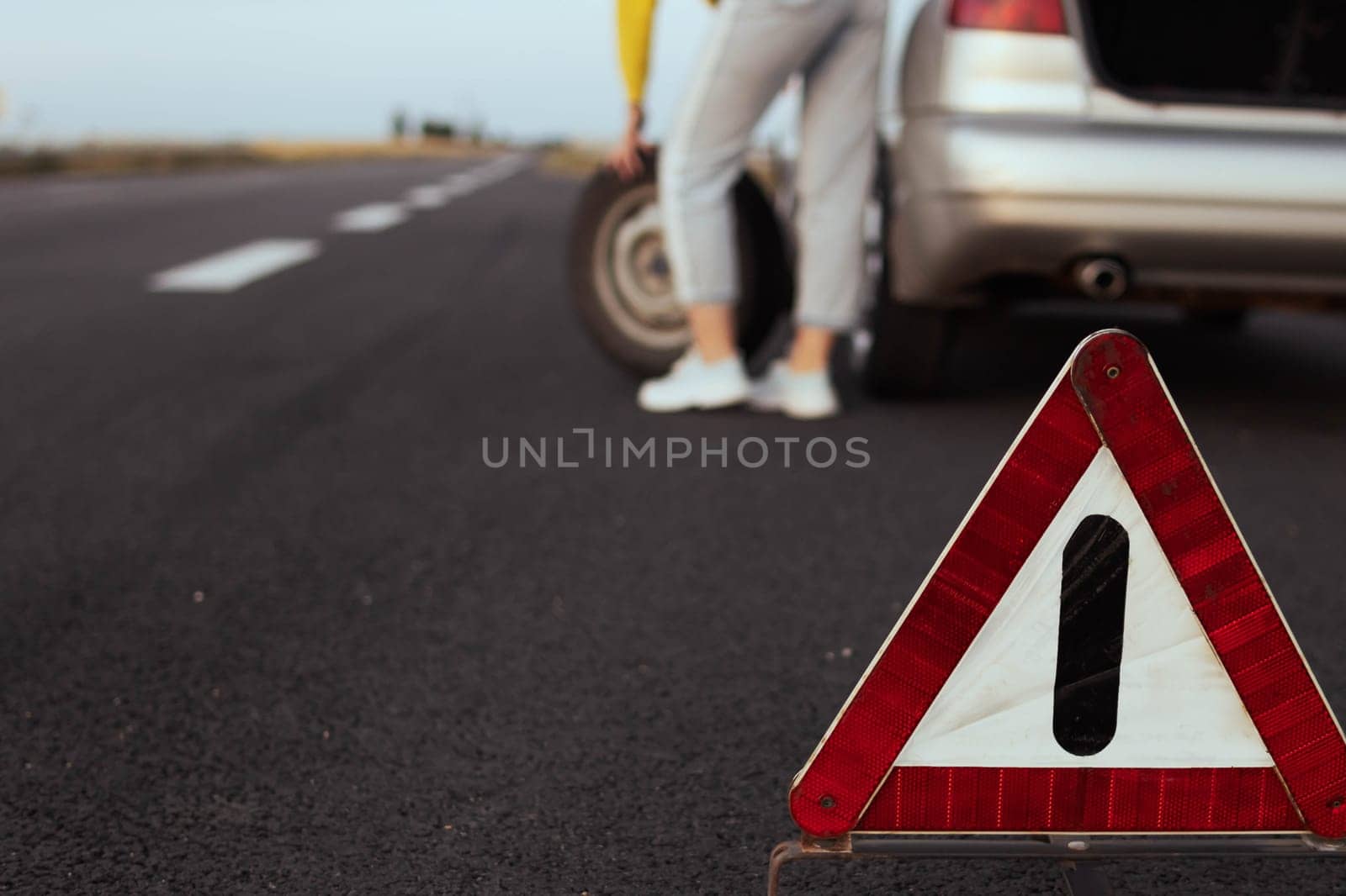 girl stands near the rear wheel of the car and rolls the spare wheel to change, the wheel has been pierced. Close-up and focus on the emergency stop sign, the background is blurred. High quality photo