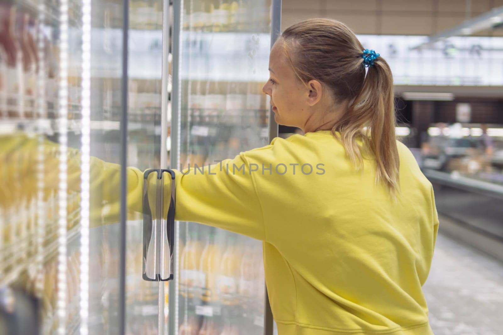 a girl with blond hair in a ponytail in a yellow sweater with long sleeves, in a grocery store, chooses products in a refrigerated display case. The concept of shopping for home. High quality photo