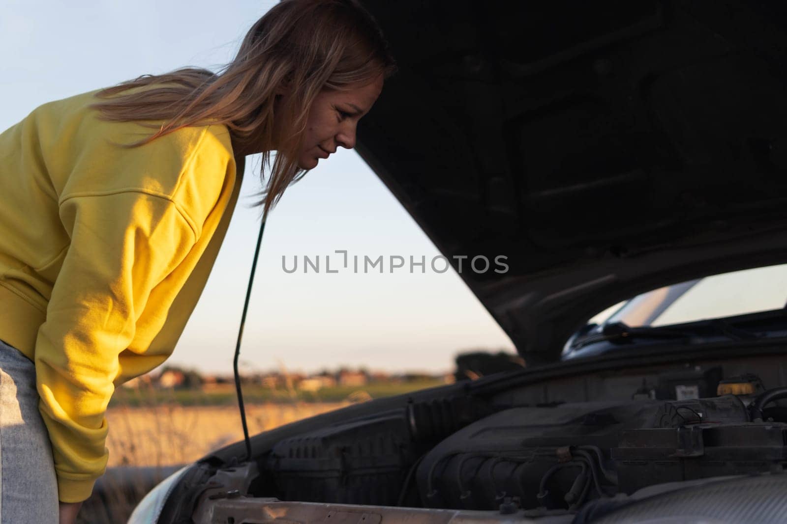 a girl with blond hair in casual clothes, stands with an open hood of a car and looks at what has broken. A car breakdown on the road. there is a place for an inscription. High quality photo
