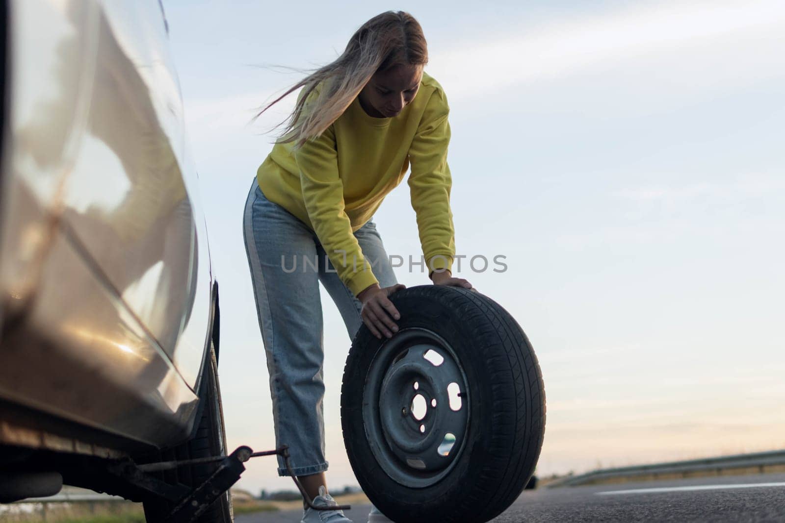 a girl in casual clothes stands near the rear wheel of the car and rolls the spare wheel to change. Car breakdown, the wheel has been pierced. Close-up by PopOff