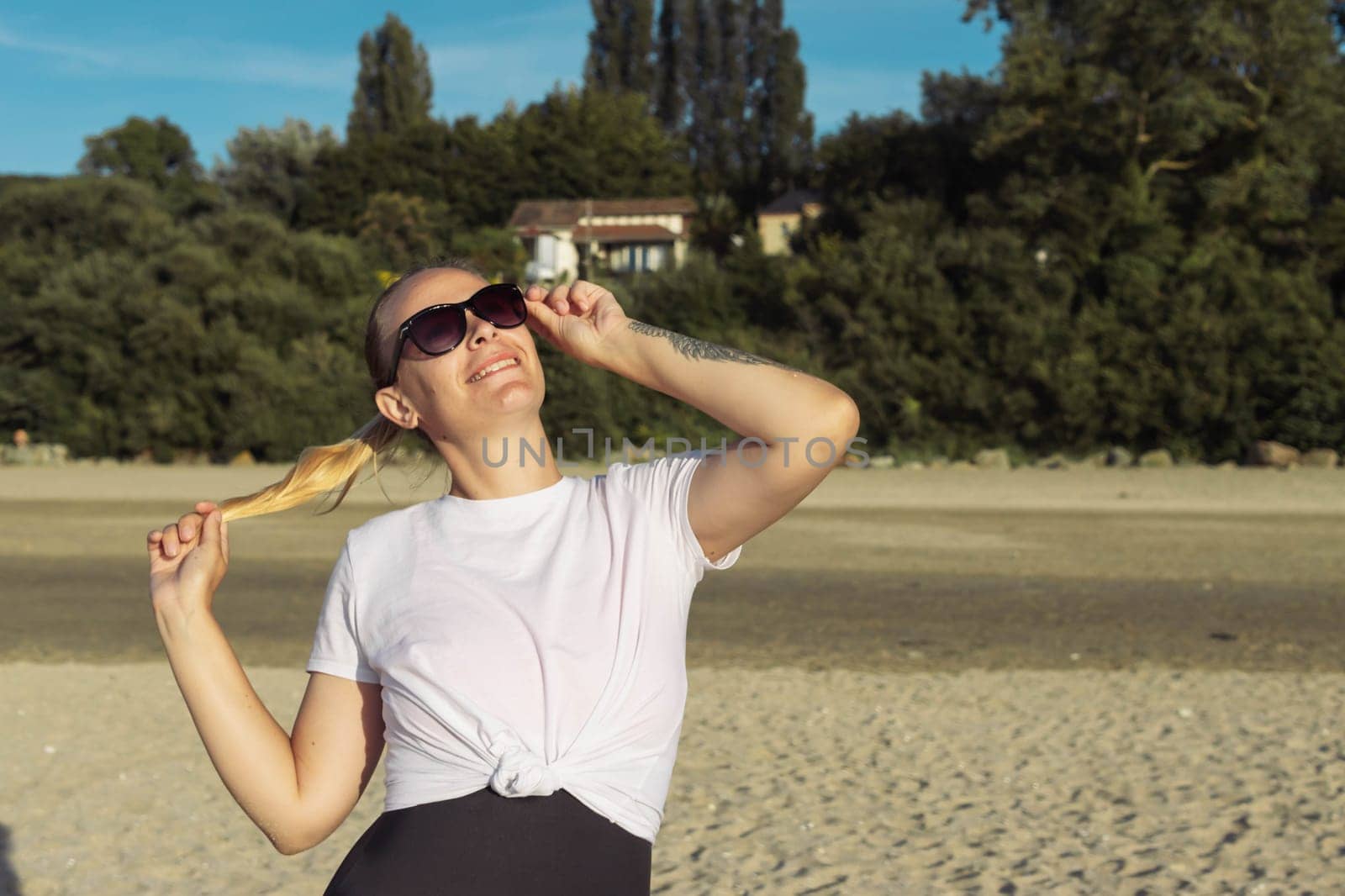 A girl with blond hair tied in a ponytail stands on the beach in a white T-shirt and swimsuit, in glasses looks up and smiles. There is a place for an inscription on the right. Beautiful landscape by PopOff