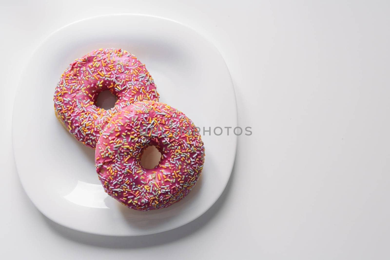 two donuts with a pink icing and colored sprinkles on a white plate lie on a white table, on the right there is a place for an inscription. High quality photo