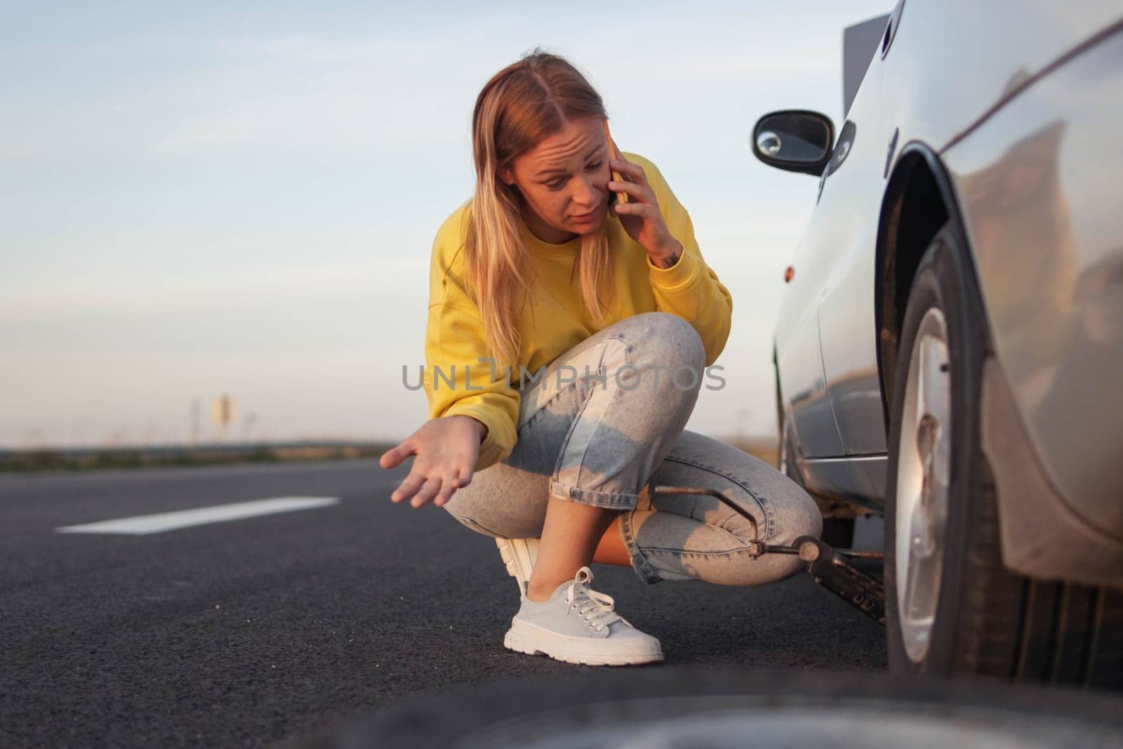 a girl in a yellow sweater and jeans of European appearance sits near the rear wheel of a punctured one, speaks on the phone how to make a wheel. With a surprised and incomprehensible face what to do by PopOff