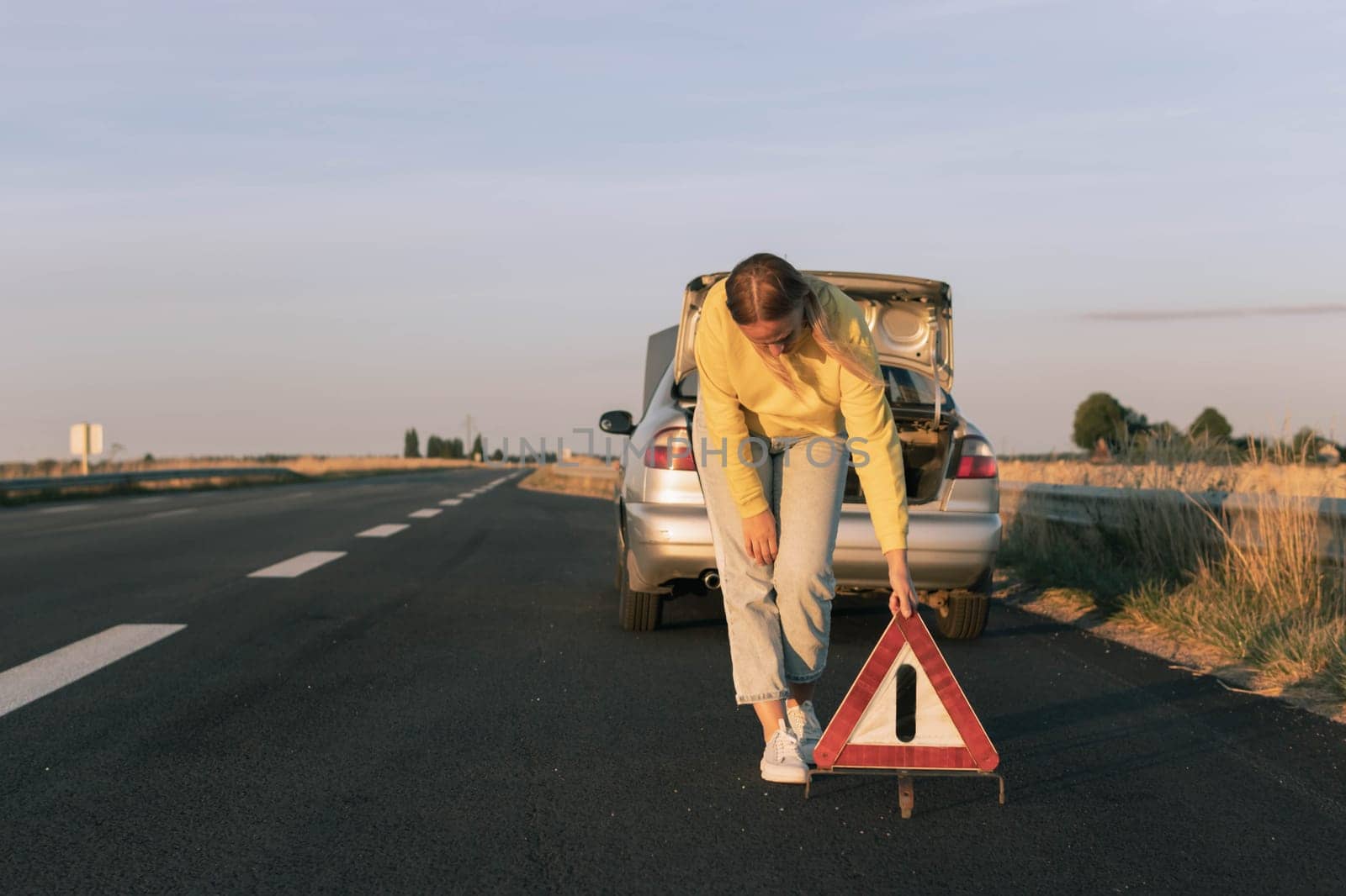 a girl with blond hair in a yellow sweater and jeans puts an emergency stop sign on the highway near a broken car. Car breakdown on the road by PopOff