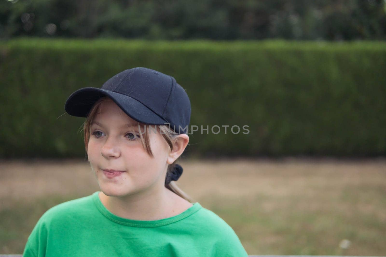 a teenage girl of European appearance was dreaming in a black cap, in a green T-shirt, standing in a park around green grass on the right there is a place for an inscription by PopOff