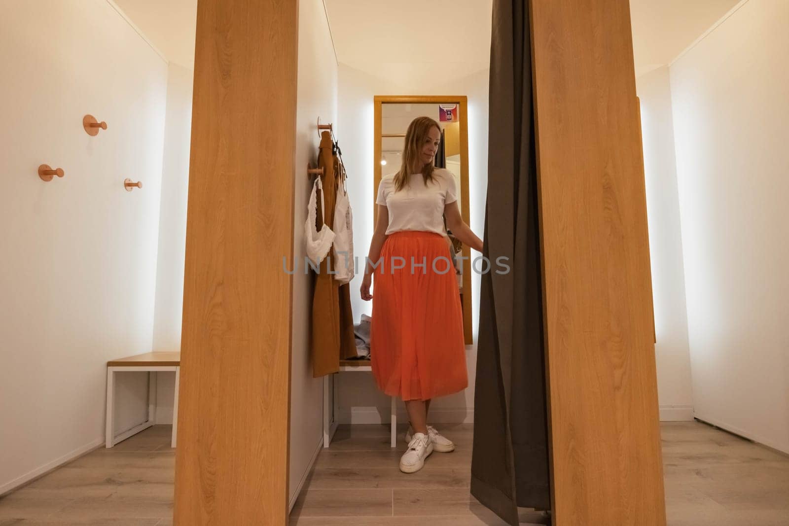 a girl trying on a white t-shirt and an orange skirt in a booth looks in the mirror. brown and white fitting booths in a things store. there is a place for an inscription. High quality photo
