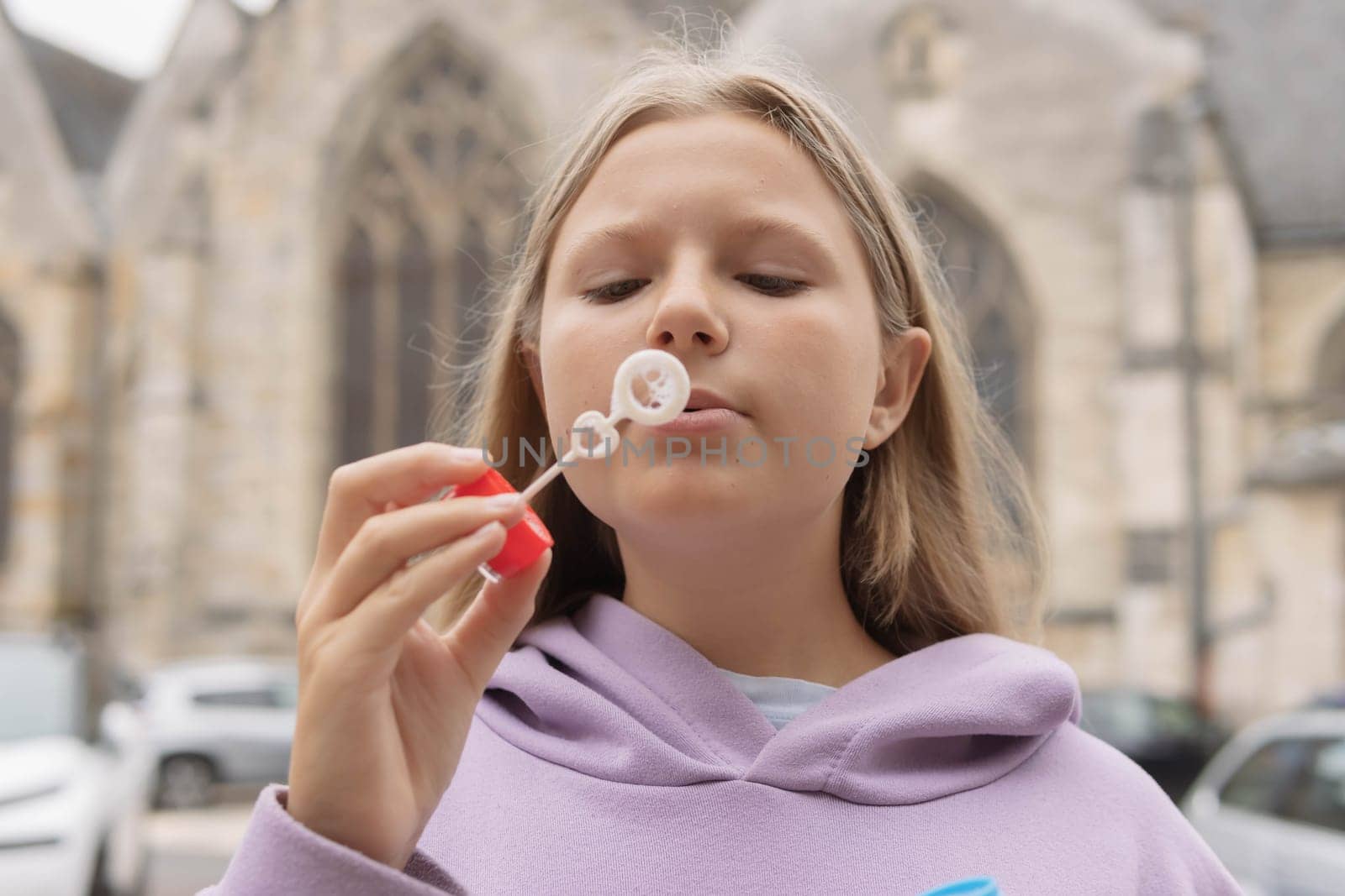 a girl of European appearance with blond hair in a lilac sweater plays with bubbles on the street. High quality photo