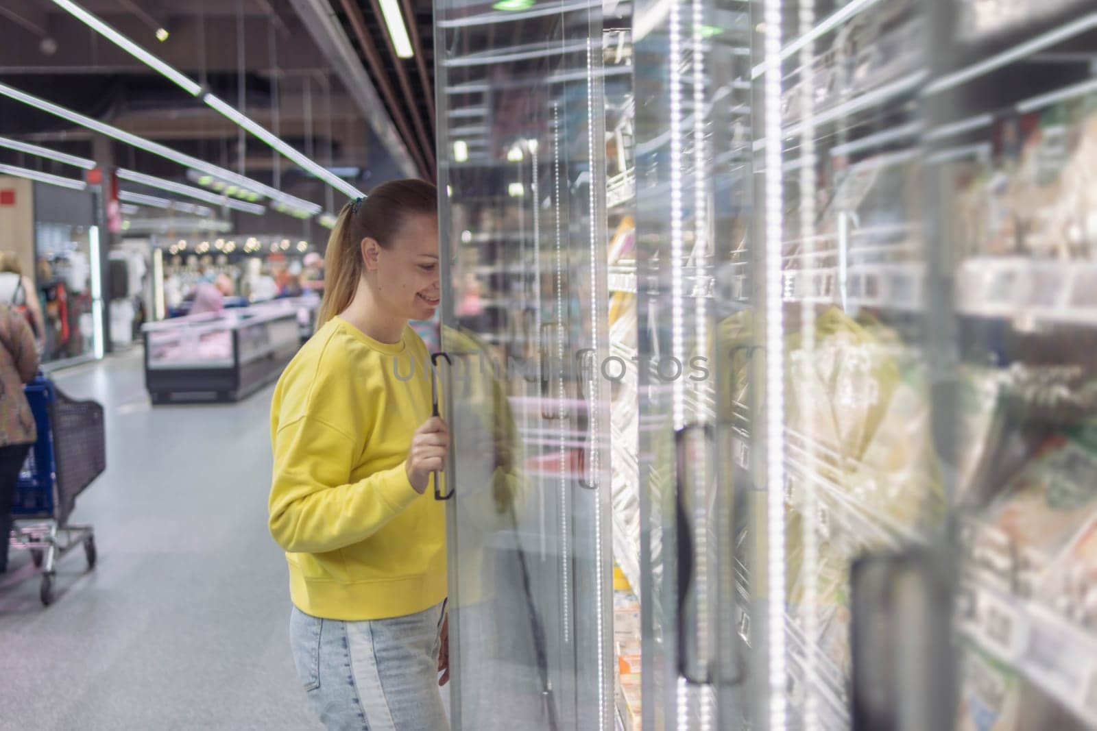 a woman of European appearance with blond hair tied in a ponytail in a yellow sweater and jeans, in a store in a refrigerated display case chooses products for the home. The concept of home food by PopOff