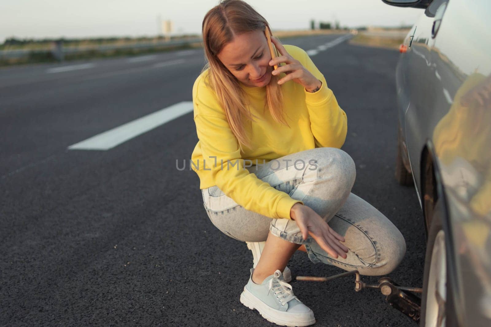 a girl in a yellow sweater and jeans of European appearance sits near the rear punctured wheel in her hands holds a key for the wheels and speaks on the phone how to make a wheel by PopOff