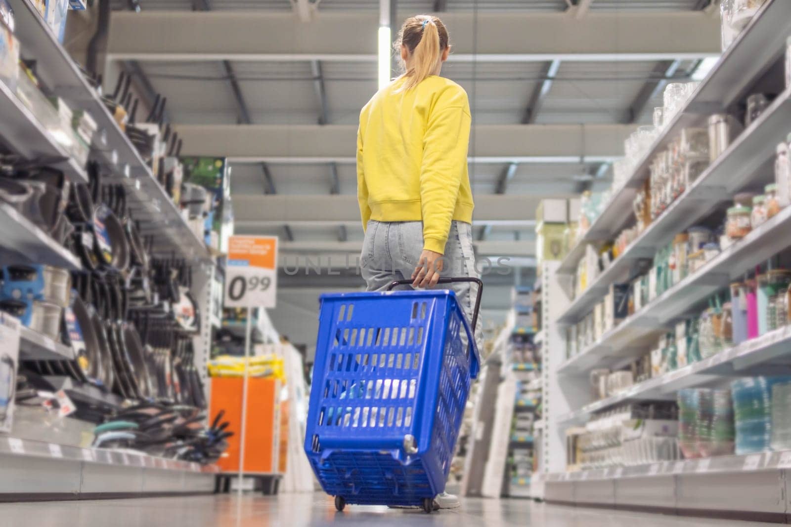 a girl with blond hair tied in a ponytail in jeans and a yellow sweater stands between the counters in a store with a blue plastic cart turned her back. High quality photo