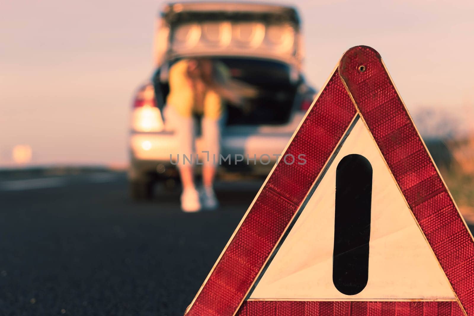 girl sits in the trunk holding her head, in the foreground is an emergency stop sign close-up and in focus, the girl is blurred in the background. Car breakdown on the highway, on the road by PopOff