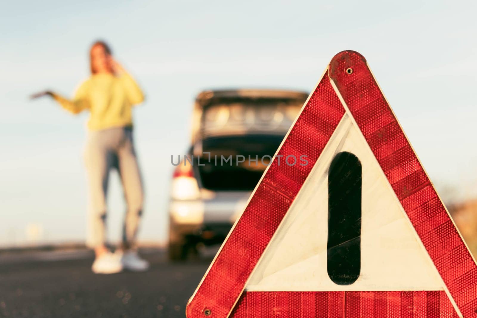 in the foreground is a red-white close-up warning triangle, in the background a girl stands with her hand raised, speaks on the phone, stops the car for roadside assistance. Car breakdown on the road by PopOff