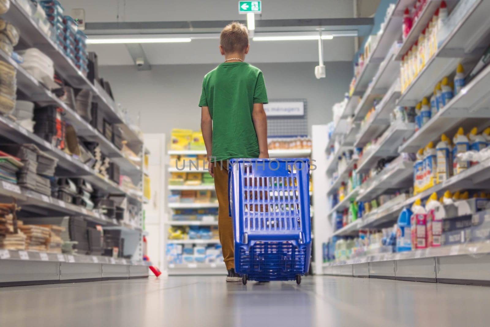 a boy of European appearance with blond short hair in a green T-shirt in a store with his back turned with a blue shopping cart, walks and chooses toys by PopOff