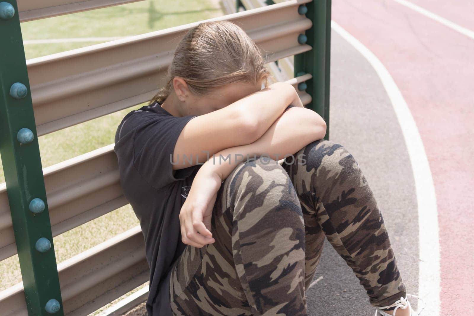 a teenage girl with blond hair sits by a fence near a football field, covering her face with her hands. in white sneakers, a dark T-shirt and green leggings. High quality photo