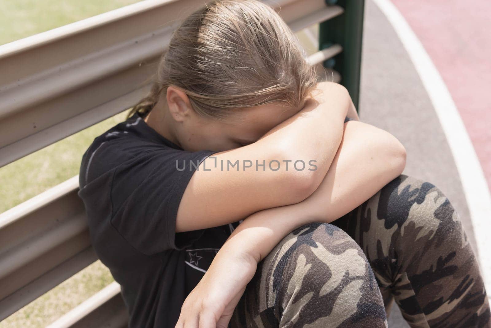 a teenage girl with blond hair sits by a fence near a football field close up by PopOff