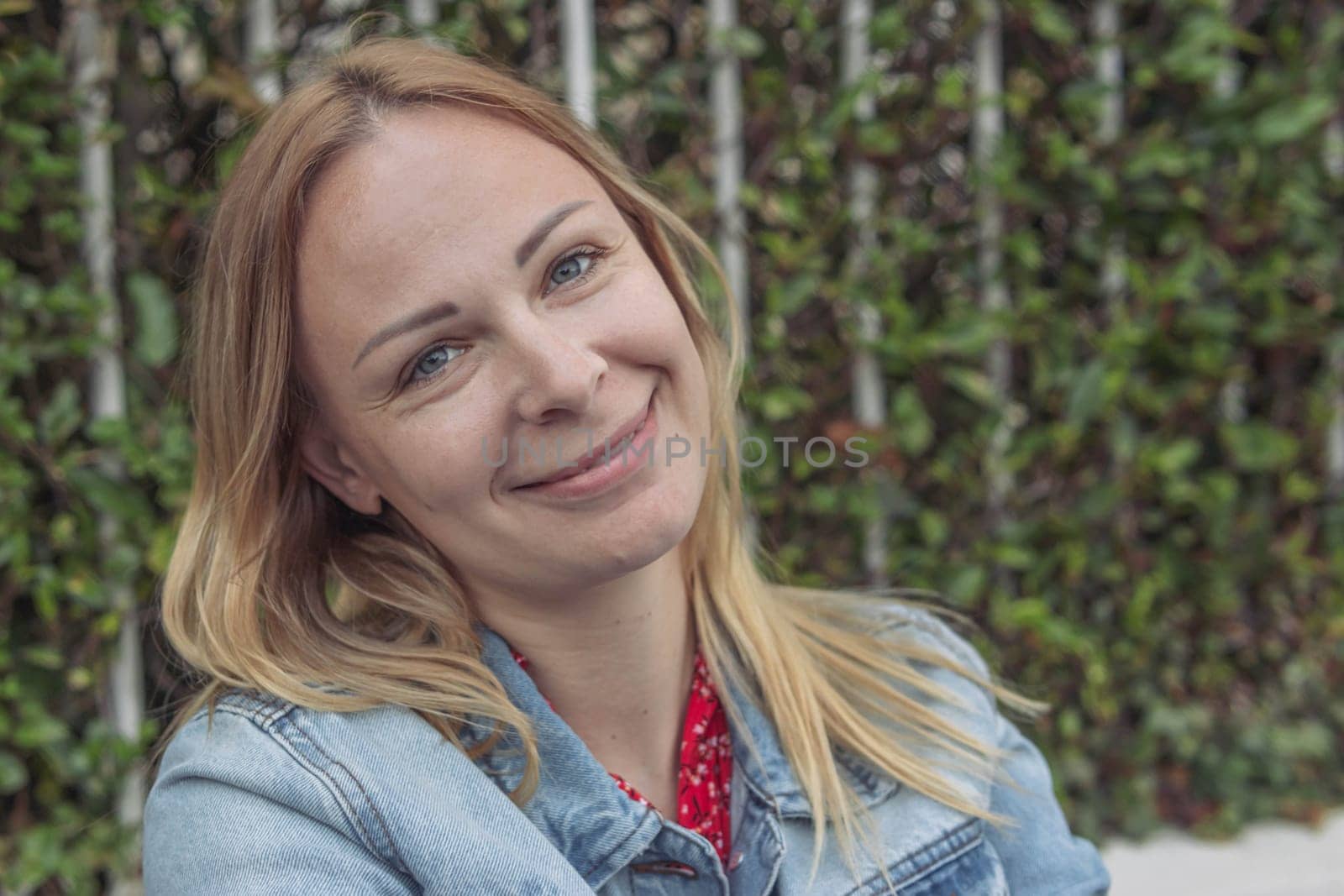 a girl with blond hair in a denim jacket sits in a park dreaming and smiling close-up, portrait .human emotions by PopOff