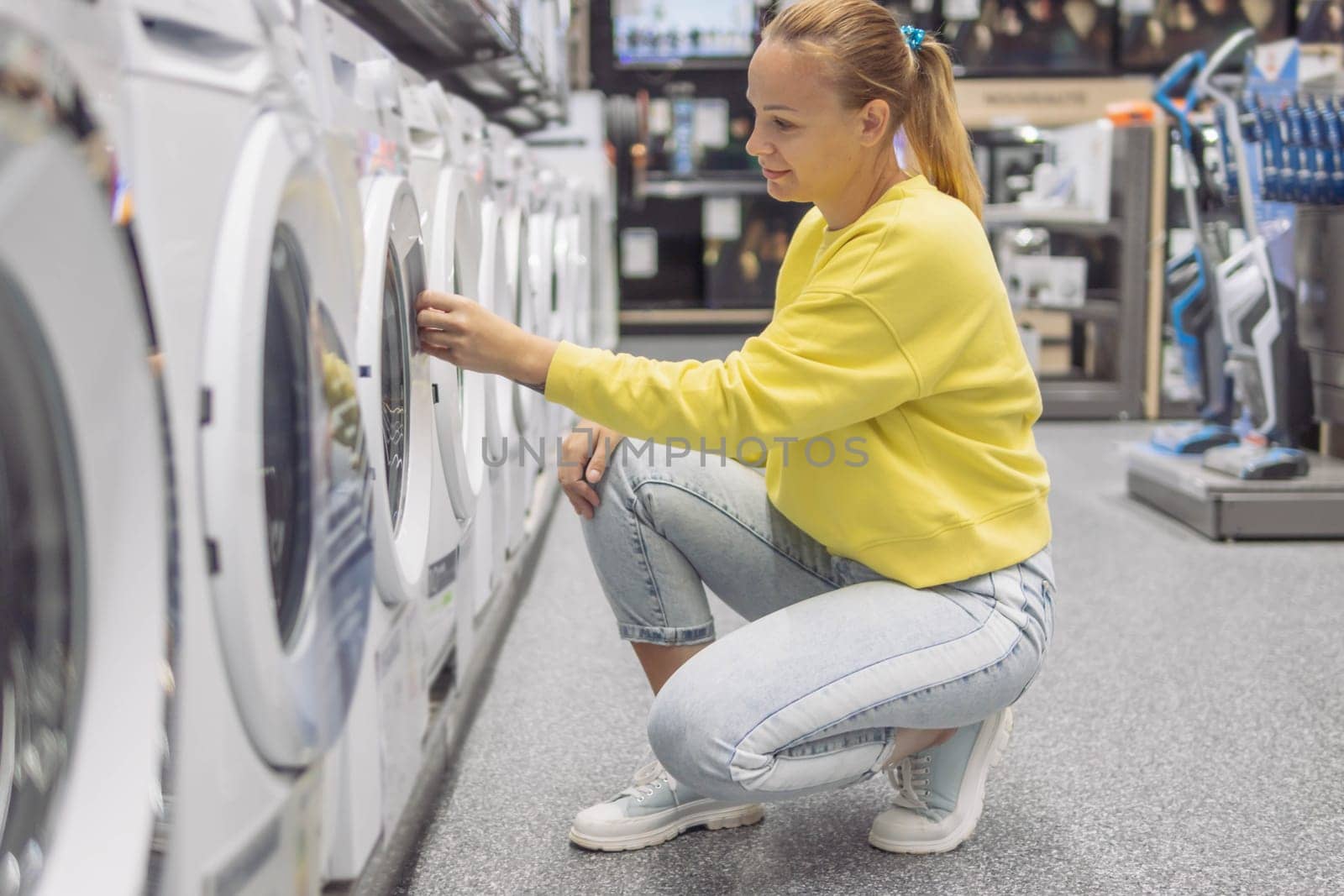 a girl of fair appearance with blond hair tied in a ponytail in a yellow sweater in jeans and light sneakers, in the store she is choosing a washing machine. High quality photo