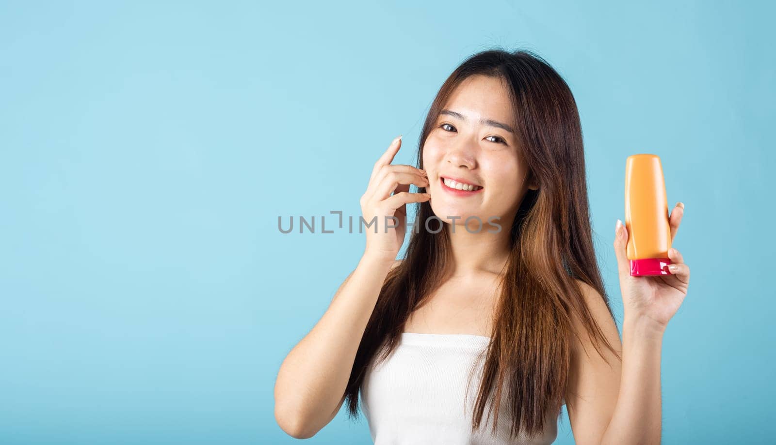 Happy smiling teen female present sun protection skin care studio shot isolated on blue background, Asian young woman holding sunscreen lotion cream with hand touching on face