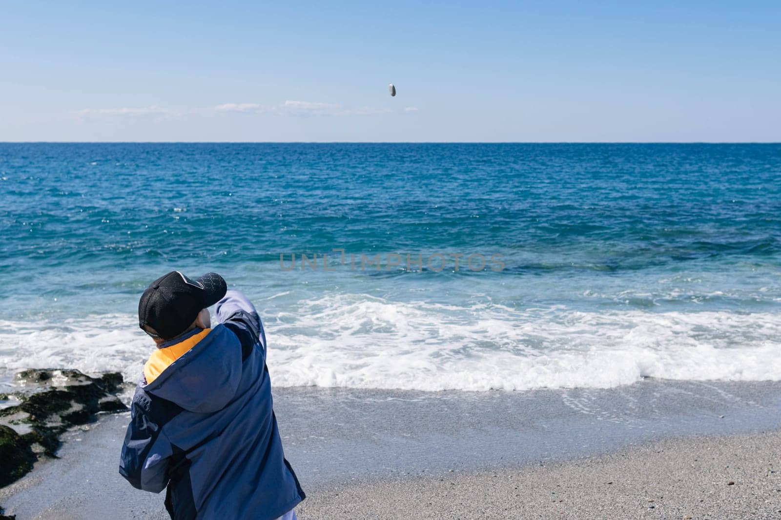 a child stands on the seashore and throws pebbles into the sea. by PopOff