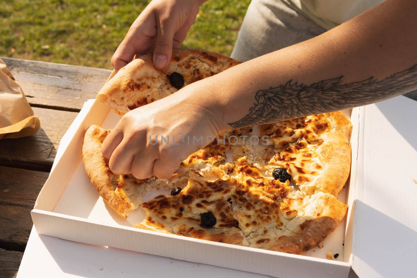 a girl with a tattoo on her arm in the shape of a wing breaks pizza at a picnic in the park, hands and pizza close-up. High quality photo