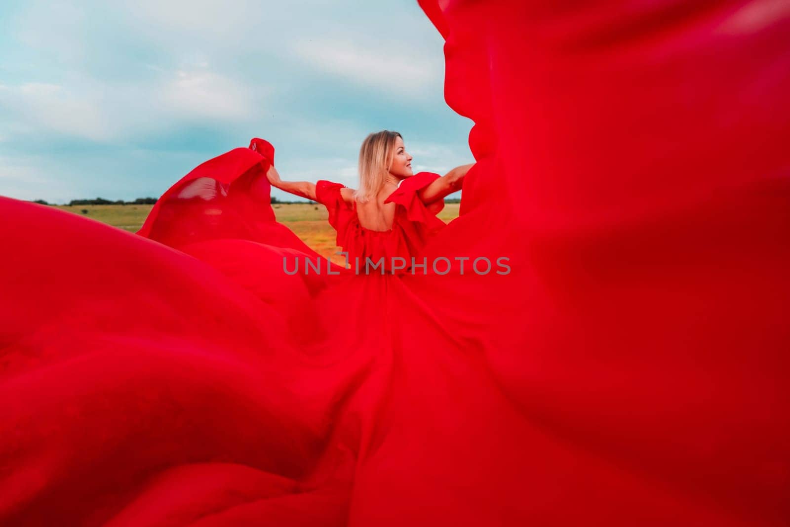 Woman Red Flying Dress. A blonde in a red dress against the sky. Rear view of a beautiful blonde woman in a red dress fluttering in the wind against a blue sky and clouds. by Matiunina