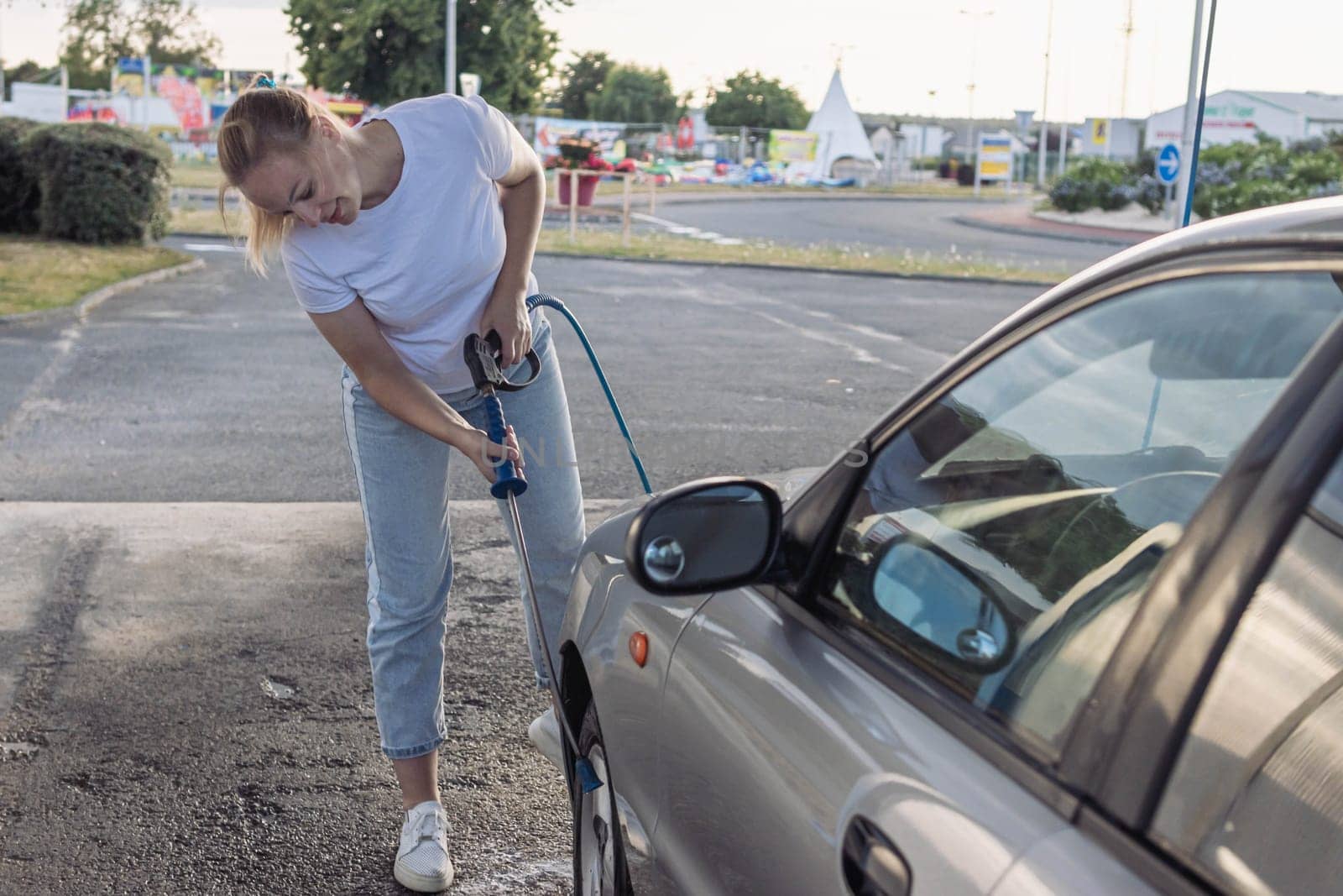 a cheerful girl in a white t-shirt with blond hair tied in a ponytail washes a gray car with water outdoors.The concept of work by PopOff