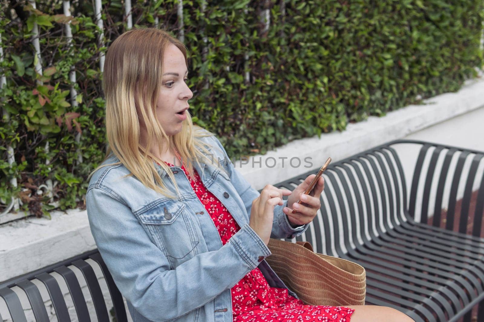 a girl of European appearance with blond hair sits on a bench and looks at the phone in surprise. The girl is dressed in a red dress and a denim jacket by PopOff