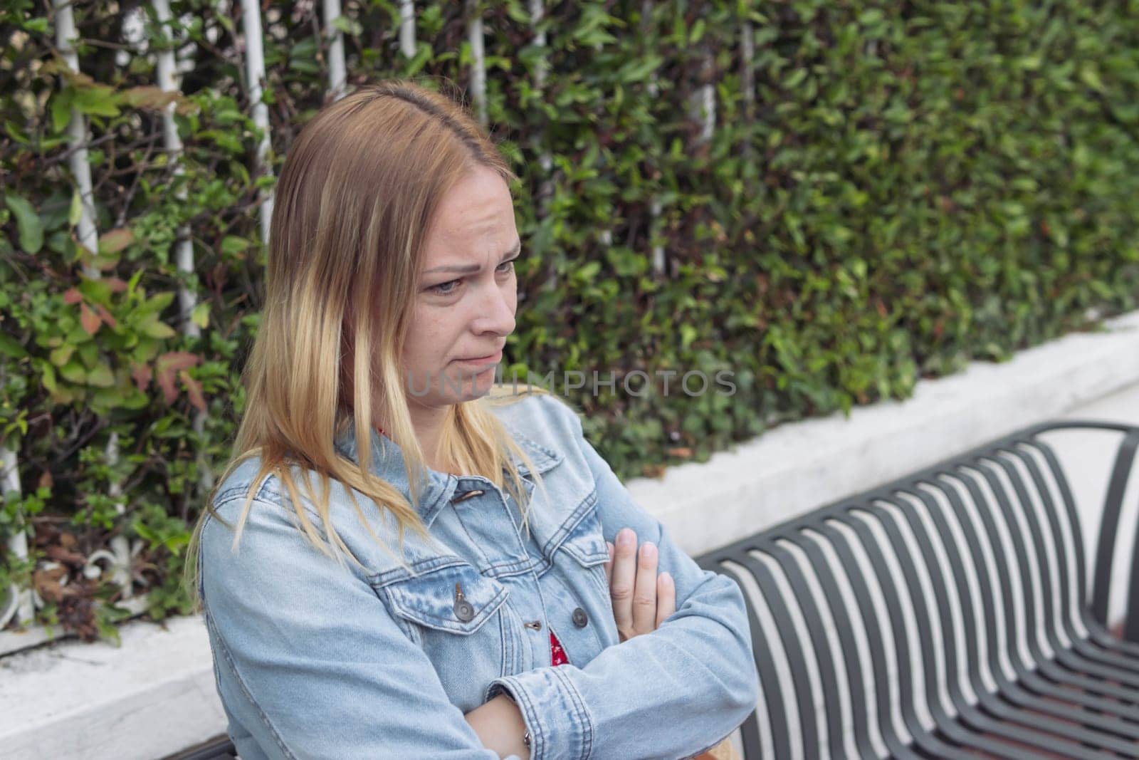 a girl of European appearance with blond hair in a red dress, a denim jacket sits on a bench offended. Human emotions. High quality photo