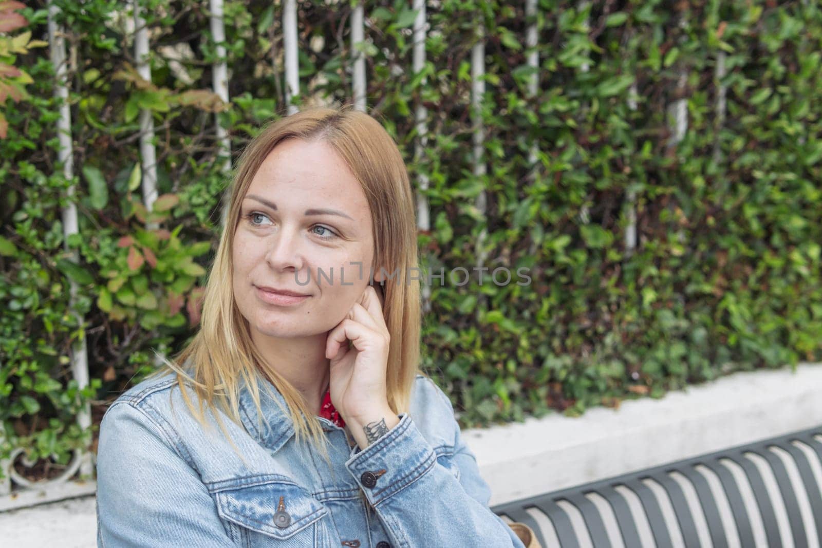 a girl of European appearance with blond hair in a denim jacket sits in a park and thinks daydreams holding her hand to her head close-up portrait on the right there is a place for an inscription. by PopOff