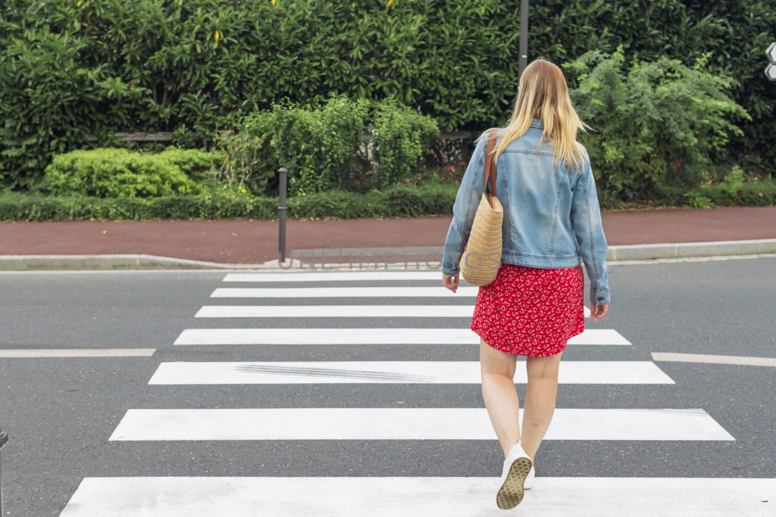 a girl of European appearance in a red dress, a denim jacket and with a bag on her shoulder crosses the road at a pedestrian crossing rear view from the front beautiful green trees. High quality photo