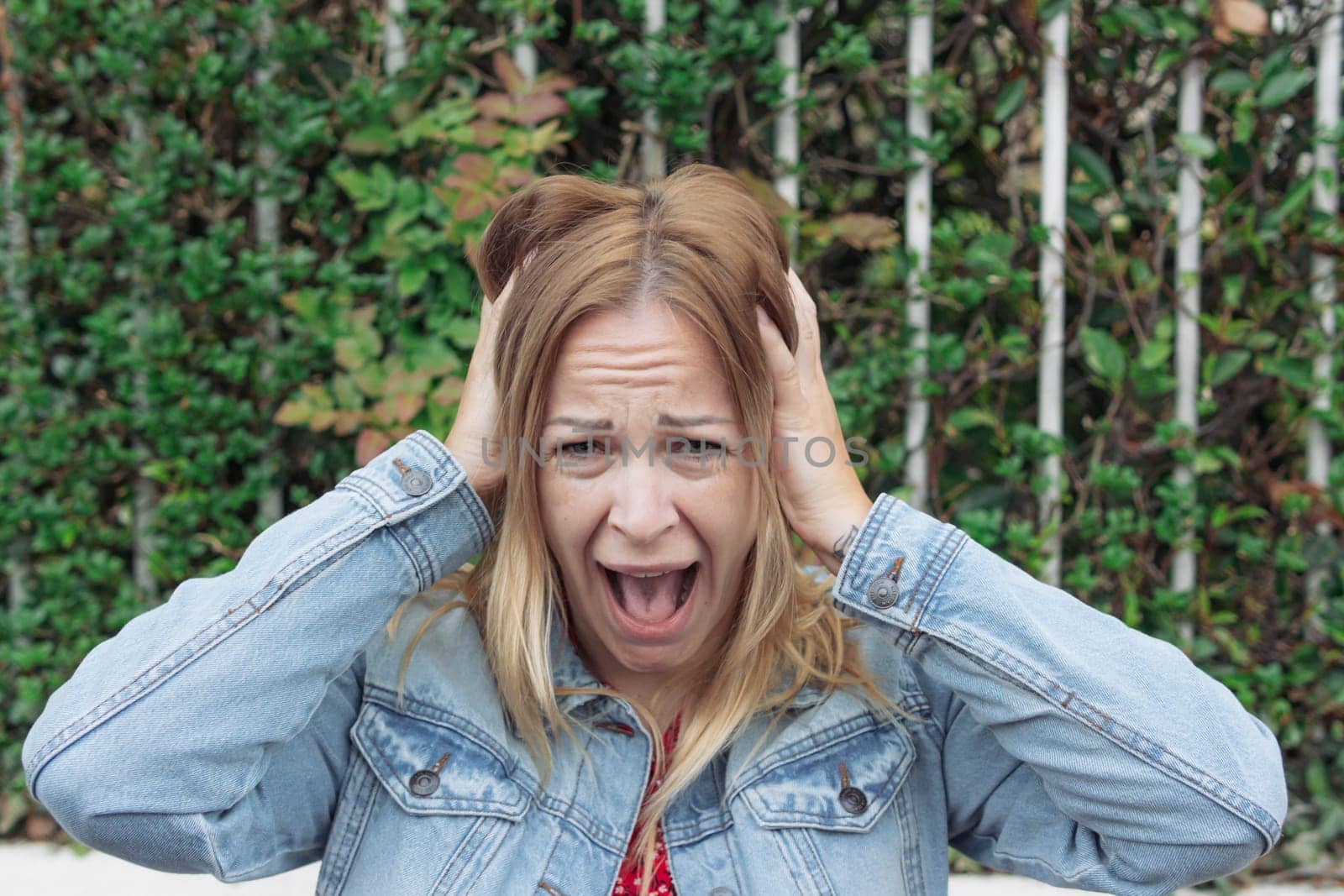 a girl of European appearance with blond hair in a denim jacket sits in a park and screams holding her head close-up. Human emotions by PopOff