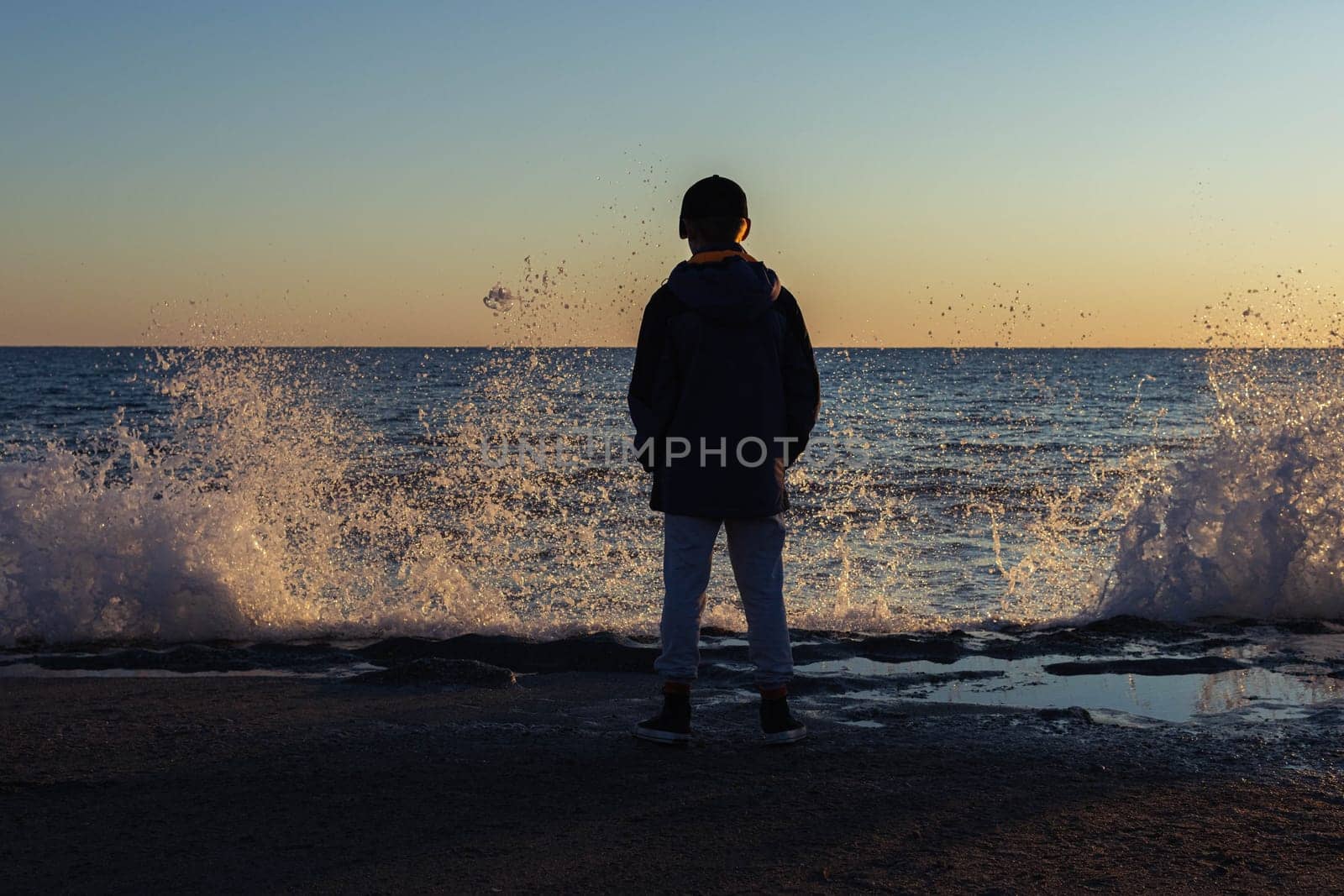 the child is standing on the seashore, photo from the back, the waves are raging at sunset, the background. High quality photo