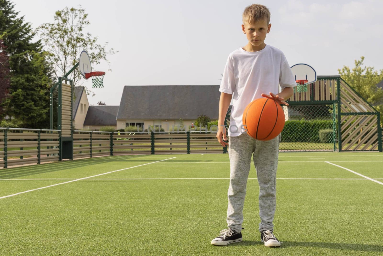 a boy in a white t-shirt and gray pants stands on a basketball court with an orange basketball by PopOff