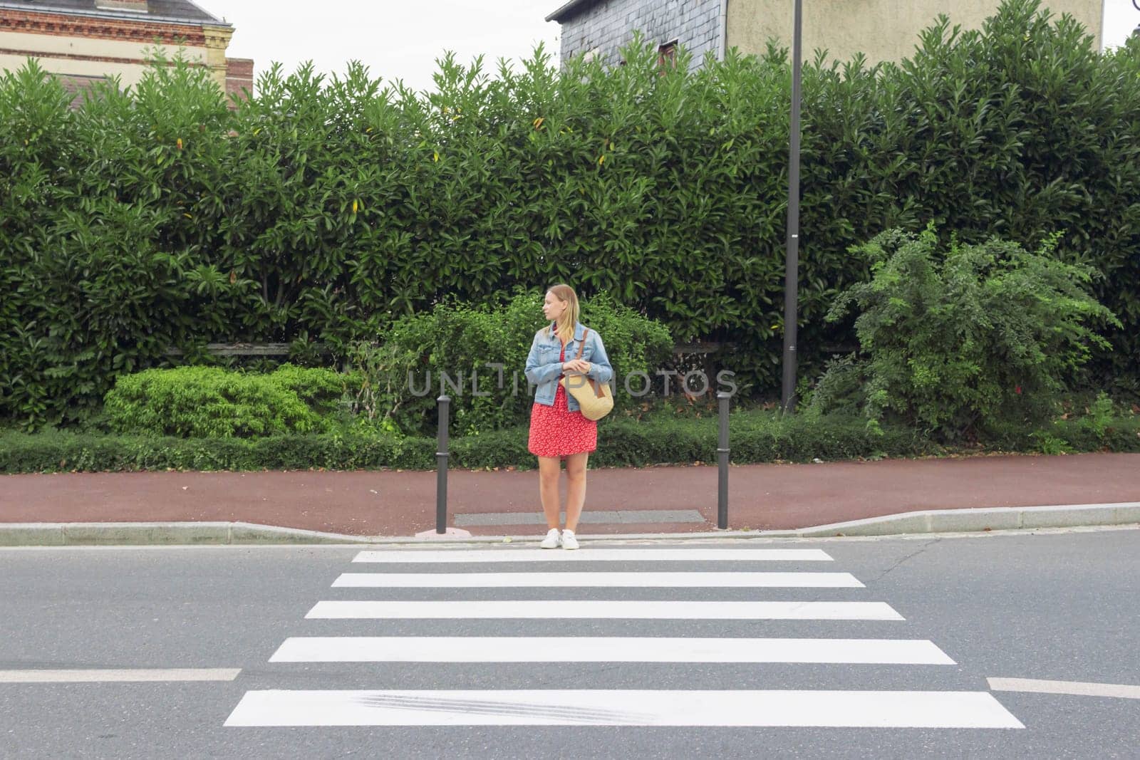 A girl in a red dress and a denim jacket with a bag on her shoulder stands at a pedestrian crossing and looks to cross the road by PopOff