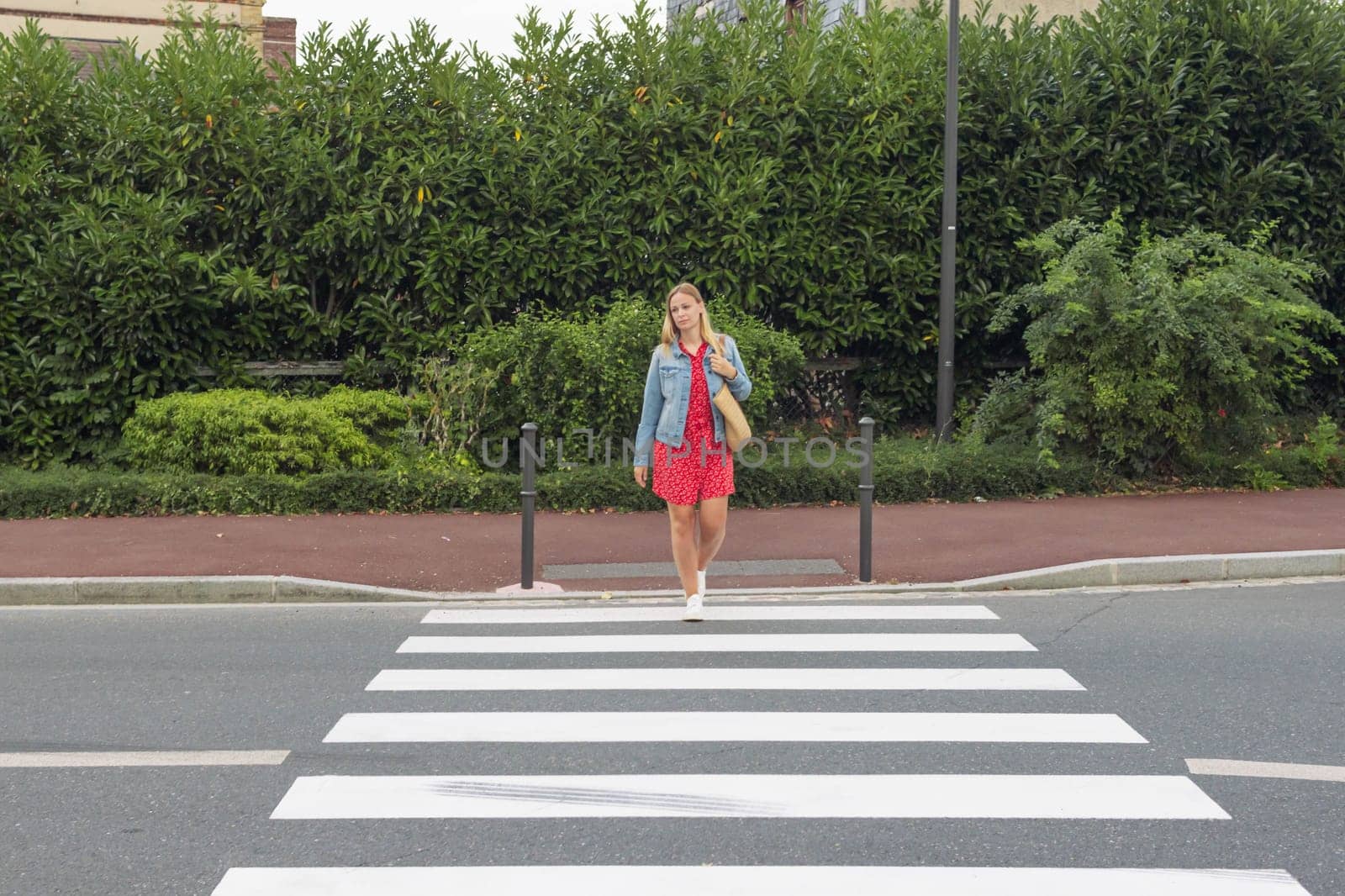 A girl with fair hair in a red dress and a denim jacket with a bag on her shoulder crosses the road at a pedestrian crossing.a pedestrian crossing on the road for safety. High quality photo