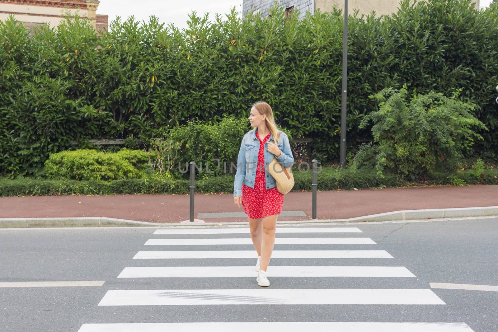 A girl in a red dress and a denim jacket with a bag on her shoulder stands at a pedestrian crossing and looks to cross the road by PopOff