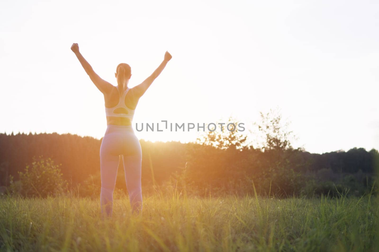 a girl in a fitness suit stands with her back to the camera at sunset with her hands raised after by PopOff