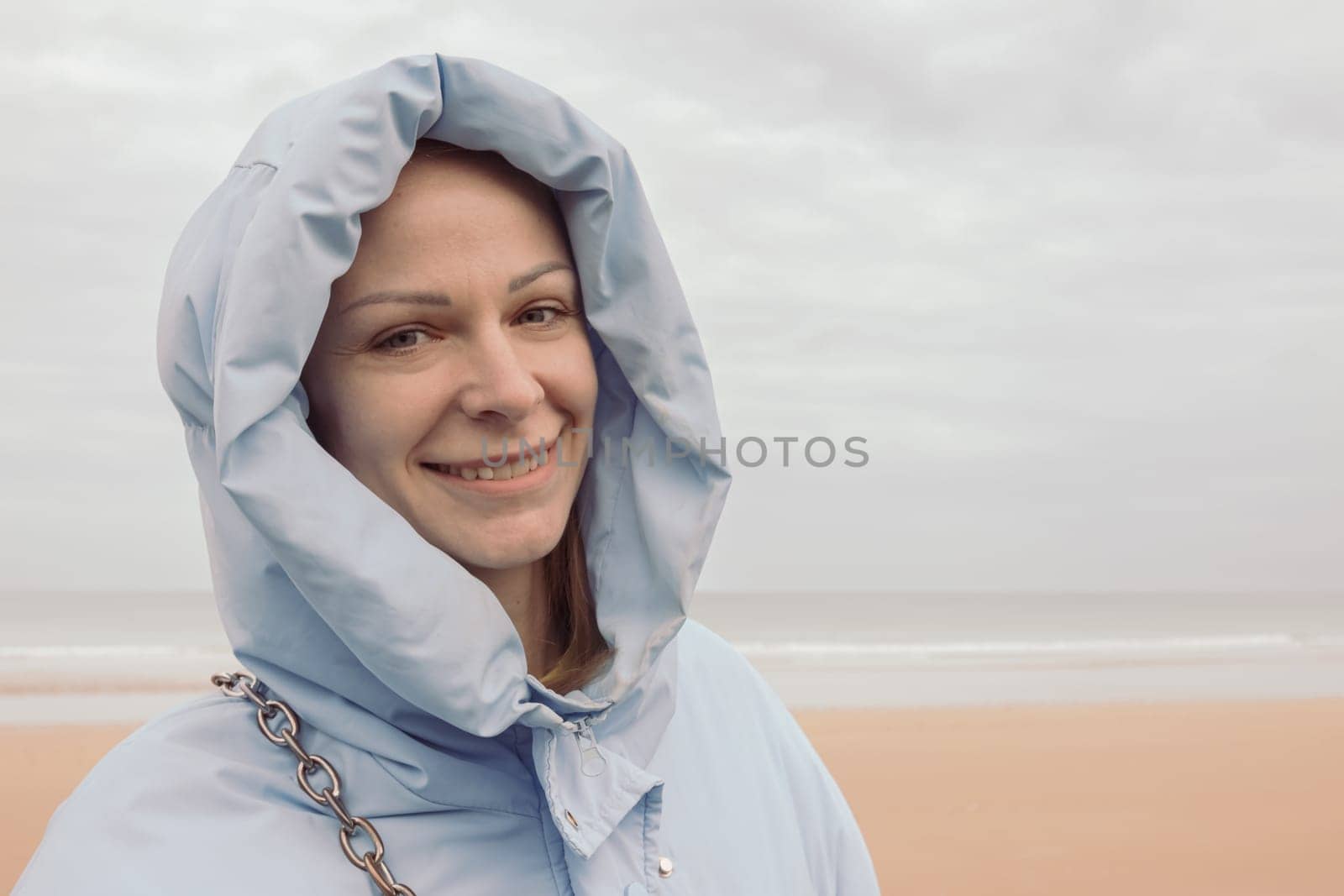 a girl of European appearance stands on the seashore in a blue jacket in a hood and smiles, looks into the camera by PopOff