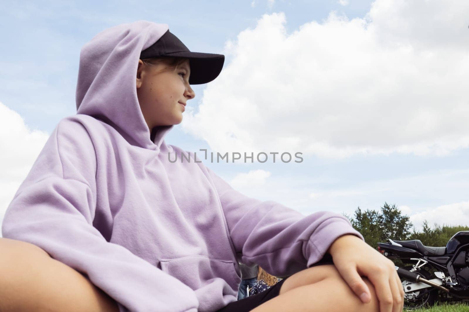 a teenager girl of European appearance in a black cap and a purple sweatshirt sits on the grass by PopOff