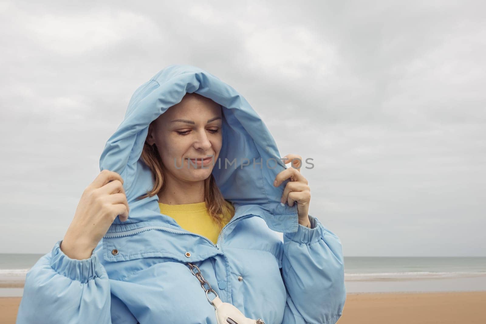 a girl of European appearance with blond hair stands on the seashore in a blue jacket and smiles with her eyes closed by PopOff