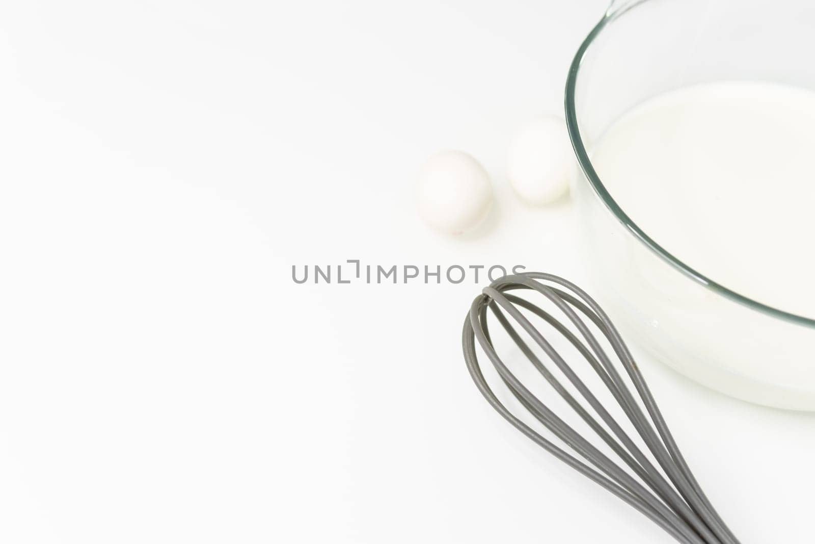 a glass transparent bowl with milk on a white table, next to a whisk for beating and eggs. close-up on a white table Culinary hobby at home - baking: cakes, muffins. High quality photo