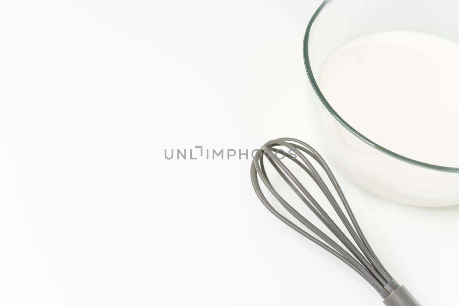 a glass transparent bowl with milk on a white table, next to a whisk for beating. close-up on a white table Culinary hobby at home - baking: cakes, muffins. High quality photo