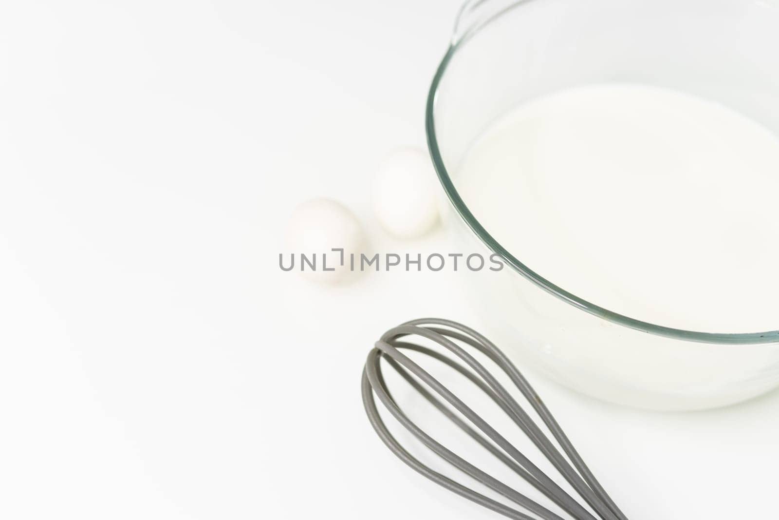 a glass transparent bowl with milk on a white table, next to a whisk by PopOff