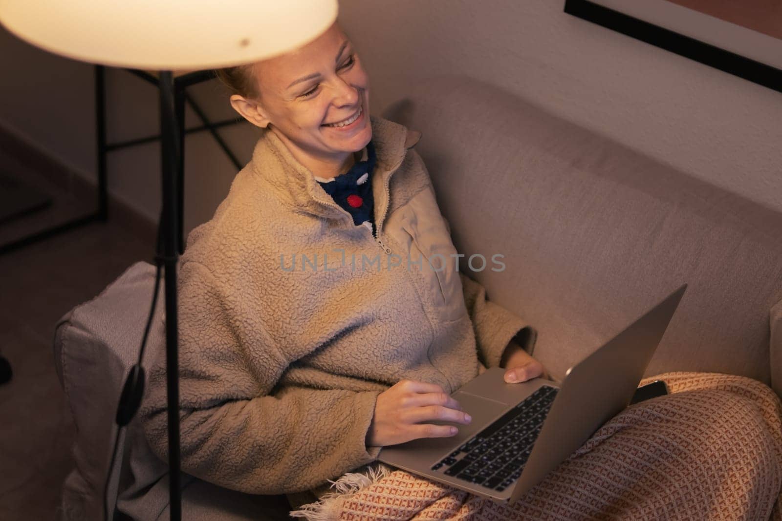 a girl of European appearance sits in the living room on the sofa covered with a brown blanket works on a laptop, the girl smiles there is a place for an inscription. High quality photo