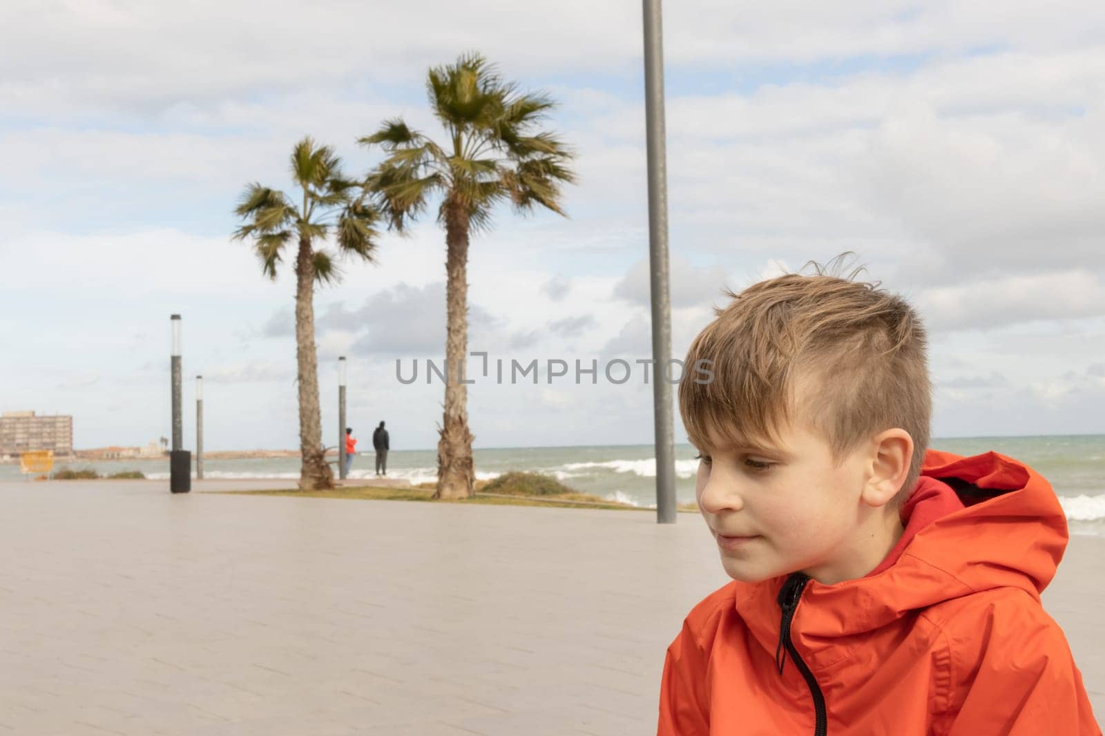 a boy of European appearance with blond hair in an orange windbreaker, standing on the embankment close-up, there is a place for an inscription by PopOff