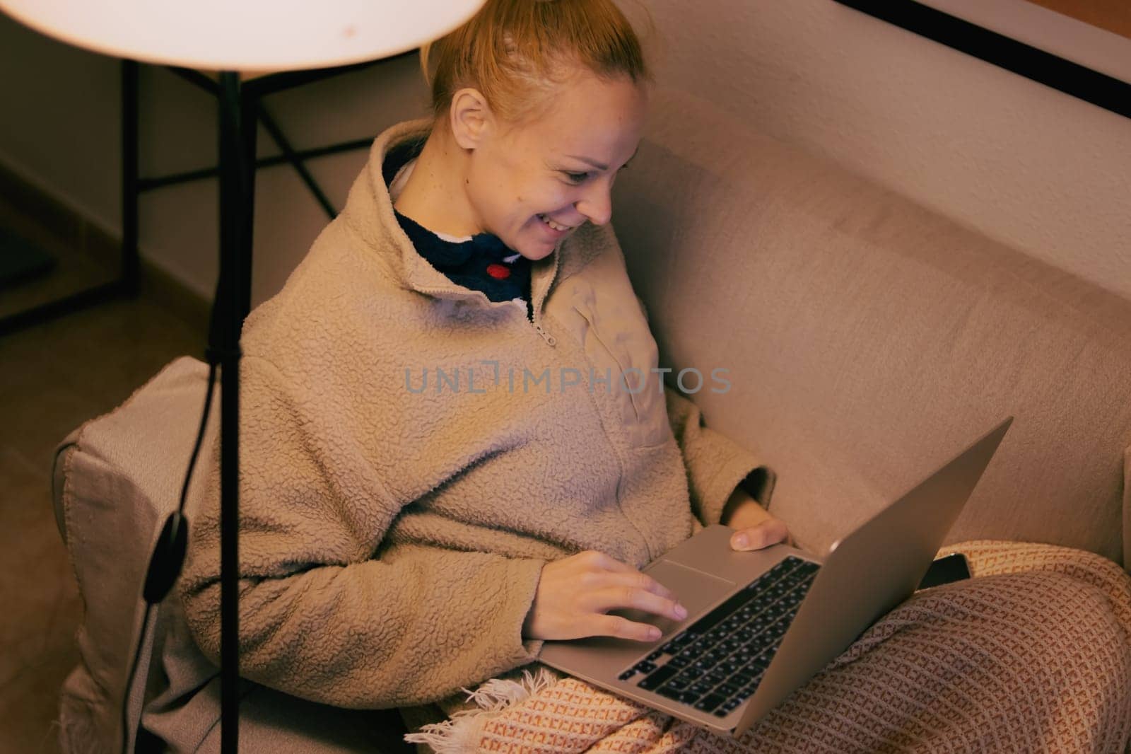 a woman of European appearance sits in the living room on the sofa covered with a brown blanket, works on a laptop, there is a place for an inscription by PopOff